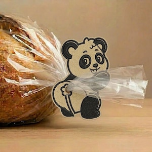 May include: A tan and black embroidered panda bear bread clip made of fabric. The panda is depicted in a seated position, holding a small flower, and is attached to a clear plastic bag containing a loaf of bread, securing the packaging closed.