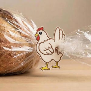 May include: A decorative chicken-shaped bread tie. The embroidered design features a white chicken with a red comb, yellow beak and feet, and brown outline. The tie is holding a clear plastic bag containing a loaf of bread.