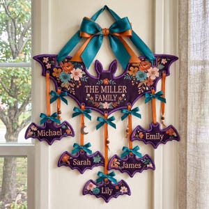 Op de afbeelding: Een decoratieve wandhanger in de vorm van een grote paarse vleermuis met bloemmotieven, bekroond met een blauwgroene en oranje strik. Daaronder hangen vijf kleinere vleermuisvormige ornamenten, gepersonaliseerd met de namen Michael, Sarah, James, Emily en Lily.