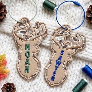 May include: Two deer-shaped luggage tags made of tan material with camouflage-patterned antlers. One tag features the name NOAH embroidered in green, and the other features the name SAWYER embroidered in blue, each attached to a circular wire loop.