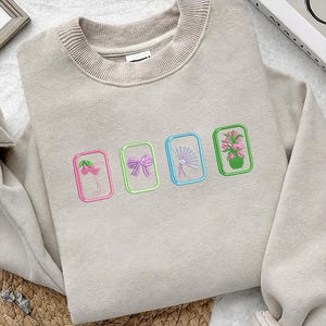 Puede incluir: Una sudadera beige de cuello redondo con cuatro parches rectangulares bordados. Los dise&ntilde;os incluyen una copa de c&oacute;ctel rosa con cerezas, un lazo morado, un abanico azul claro y un jarr&oacute;n verde lleno de flores rosas.