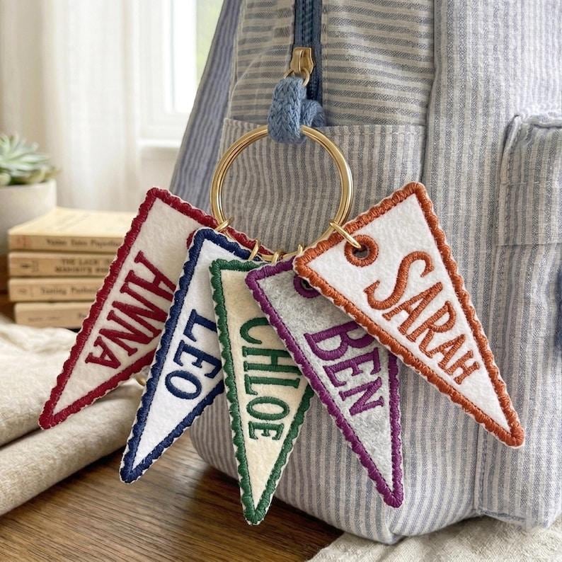 May include: A collection of personalized felt pennant keychains. Each pennant is a different color, with embroidered names in various colors. The keychains are attached to a gold ring and are hanging in front of a striped backpack.