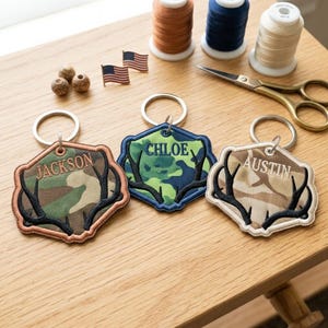 May include: Three hexagonal embroidered keychains featuring camouflage fabric, black stitched antlers, and personalised names Jackson, Chloe, and Austin. Each keychain includes a silver metal ring, with sewing supplies like thread spools and scissors nearby.