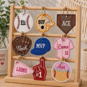 May include: A wooden display rack holding nine embroidered felt keychains with sports themes. The keychains feature shapes like baseballs, gloves, jerseys, and foam fingers, personalized with names including Eli, ACE, Noah, MVP, Emma, LIONS, and Ava.