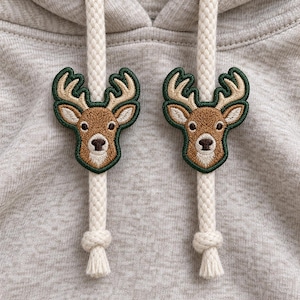May include: A light heather grey hoodie featuring two thick, cream-coloured braided drawstrings. Each drawstring is adorned with an embroidered deer head patch, showcasing a brown face, cream-coloured muzzle, and tan antlers outlined in dark green.