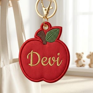 May include: A red apple-shaped keychain featuring embroidered gold text that reads Devi. The apple has a brown stem and a green leaf, attached to a gold-toned metal split ring and a lobster clasp mechanism for secure attachment to bags or keys.