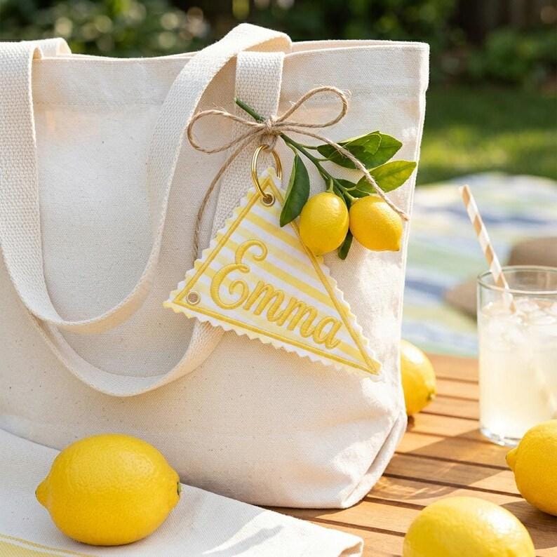 May include: Cream-colored canvas tote bag with a matching handle. A yellow and white striped tag with the name "Emma" is attached. The bag is decorated with lemons and greenery, suggesting a summer theme.