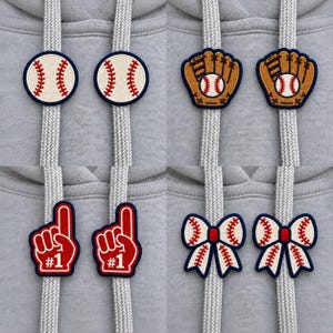 May include: Four sets of two matching embroidered drawstring charms attached to gray hoodie strings. The designs include white baseballs with red stitching, brown baseball gloves with white balls, red foam fingers with white #1 text, and white bows with red stitching.