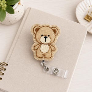 May include: A badge reel featuring a glittery tan teddy bear design with embroidered details, including a cream-colored belly and muzzle, black eyes, and a black nose. The bear is attached to a clear plastic strap with a metal snap button closure.