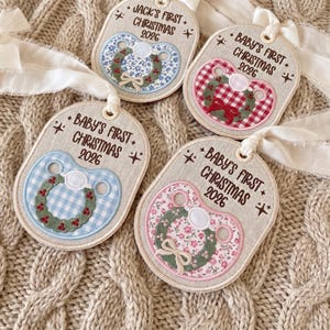 May include: Four oval-shaped fabric ornaments featuring embroidered pacifiers with floral or gingham patterns and green wreaths. Each ornament displays the text Baby's First Christmas 2026 or Jack's First Christmas 2026, accented with white ribbon ties.