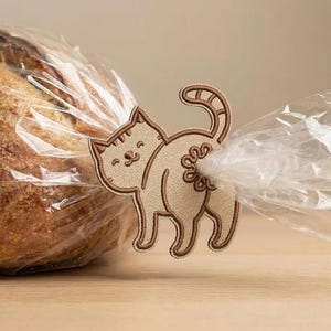 Può includere: Una clip per sacchetti a forma di gatto beige con dettagli ricamati marroni, che chiude un sacchetto di plastica trasparente. Il gatto ha una faccia sorridente e un disegno a spirale sul retro.