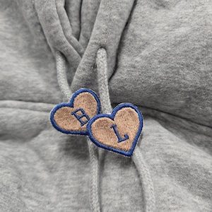 May include: A heather grey hooded sweatshirt featuring two heart-shaped patches attached to the drawstring cords. Each tan heart patch is outlined in blue embroidery and features a single blue embroidered letter, with B on one and L on the other.