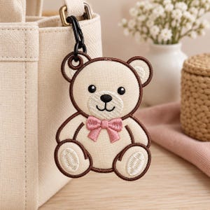 Puede incluir: Un llavero de oso de peluche bordado en color beige con un contorno cosido en marrón oscuro, ojos negros, nariz negra y una pajarita rosa. El oso está sujeto a un mosquetón de metal negro, enganchado a un bolso de lona beige.