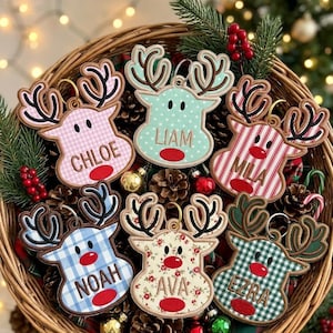 May include: Six embroidered reindeer ornaments with fabric patterns and red noses. Each ornament features a name: Chloe, Liam, Mila, Noah, Ava, and Ezra. The reindeer have brown antlers and are displayed in a woven basket with pinecones and greenery.