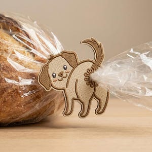 May include: A tan and brown embroidered dog-shaped bread bag clip. The clip is holding a clear plastic bag closed, with a loaf of bread visible inside. The dog has a smiling face and a curly tail.