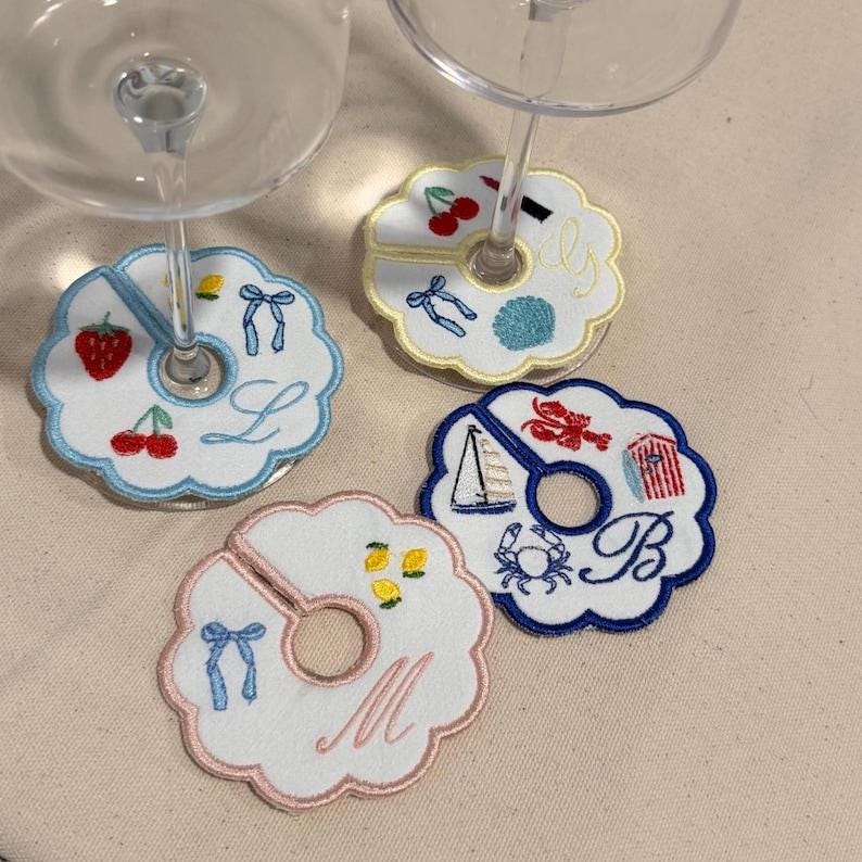 May include: Embroidered wine glass coasters with scalloped edges. Each coaster features a different design, including a strawberry, cherries, lemons, a sailboat, and a crab. The coasters have a slot for the glass stem and are decorated with letters and bows.