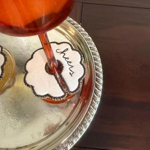May include: A silver serving tray holds a wine glass and a coaster. The coaster is white with a black embroidered border and the word "Cheers". The wine glass is filled with a reddish-orange liquid.