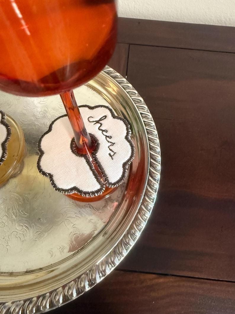 May include: A silver serving tray holds a wine glass and a coaster. The coaster is white with a black embroidered border and the word "Cheers". The wine glass is filled with a reddish-orange liquid.
