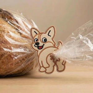 May include: A tan corgi dog-shaped bread clip, embroidered with brown thread, secures a clear plastic bag around a loaf of bread. The corgi is smiling and looking back, with its rear end facing the viewer.