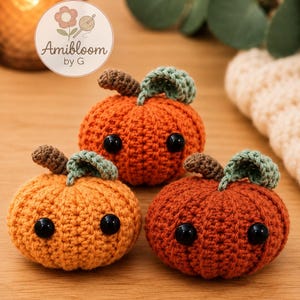 Puede incluir: Tres pequeñas calabazas de ganchillo, redondas, en tonos naranja y óxido. Cada calabaza tiene un tallo marrón, una hoja verde y dos ojos de seguridad de plástico negro. Un logotipo circular con el texto Amibloom by G es visible arriba a la izquierda.
