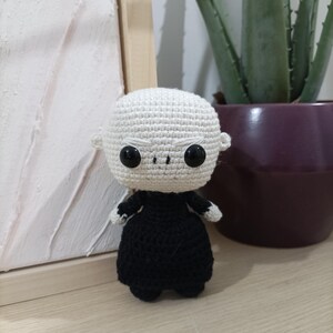 May include: A crocheted amigurumi doll with a white head and black body. The doll has large black eyes and a serious expression.