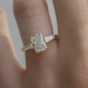 Op de afbeelding: Een gouden ring met een rechthoekige, heldere edelsteen als hoofdsteen, geflankeerd door twee kleinere, heldere, rechthoekige edelstenen. De ring is gezet op een gouden band, met een klassiek en elegant ontwerp.