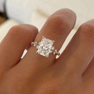 May include: A rose gold ring with a large, rectangular-cut diamond as the focal point. The ring is embellished with smaller diamonds on the band, creating a sparkling design. The ring is displayed on a finger.