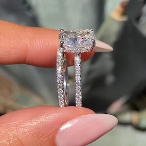 Pode incluir: Um close-up de um conjunto de anéis de diamante. O anel apresenta um grande diamante central de corte quadrado, rodeado por diamantes menores. A banda também é incrustada com pequenos diamantes, criando um efeito brilhante.