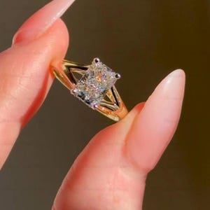 May include: A gold ring featuring a rectangular, clear gemstone. The ring has a split shank design with a triangular accent on each side of the gemstone. The gemstone is secured by four prongs.