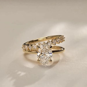 Op de afbeelding: Een gouden verlovingsring met een grote ovaal geslepen diamant als middelpunt. De band heeft kleinere diamanten langs de bovenste helft. De ring staat tegen een zachte, neutrale achtergrond, waardoor de schittering wordt benadrukt.