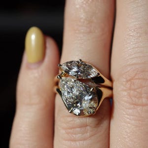 May include: Two gold rings, each with a pear-cut diamond. One ring has a single, large diamond, while the other has two smaller diamonds. The rings are stacked, and the diamonds glisten against the gold band.