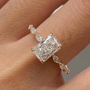 May include: A gold ring featuring a large, rectangular-cut diamond as the centerpiece. The band is adorned with smaller, marquise-cut diamonds. The ring is set in a gold band, creating a classic and elegant design.