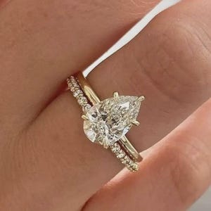 May include: A gold engagement ring with a pear-shaped diamond and a smaller band with small, round stones. The ring is set on a finger, showcasing the intricate details of the jewelry.