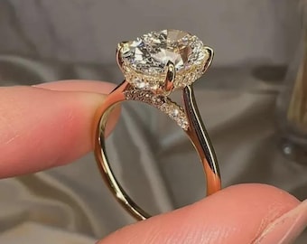 May include: A gold engagement ring with a large, clear, oval-cut diamond as the centerpiece. The ring features a halo of smaller diamonds around the main stone and along the band, creating a sparkling effect. The ring is held up by a person's fingers.