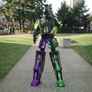 May include: A futuristic robot costume in black and green. The costume includes a helmet, chest plate, and leg armour with glowing green, purple, and pink lights. The robot is standing on a paved walkway in a park, surrounded by trees.