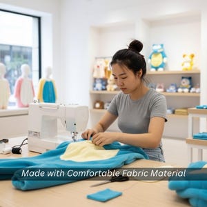 May include: A woman sewing a blue and yellow plush garment with a white sewing machine. The text "Made with Comfortable Fabric Material" is displayed at the bottom of the image. Other plush toys are visible in the background.
