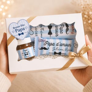 Peut inclure: Une boîte cadeau blanche avec fenêtre, attachée avec un ruban doré, contenant un pot et des barres chocolatées. Le pot et les barres ont des étiquettes à rayures bleues et blanches avec du texte et des motifs de nœuds papillon. Une étiquette en forme de cœur dit "Bonne fête Papa".
