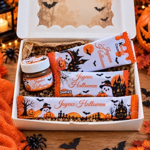 Peut inclure: Une boîte cadeau blanche remplie de papier kraft froissé contenant un petit pot de pâte à tartiner et trois barres chocolatées, avec des emballages sur le thème d'Halloween illustrés de fantômes, citrouilles, chauves-souris et Joyeux Halloween.