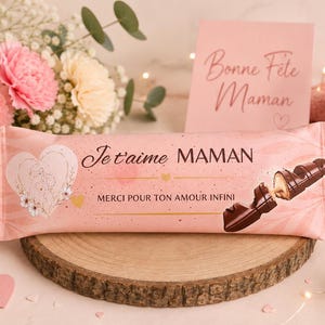 Peut inclure: Une barre de chocolat rose avec les mots "Je t'aime Maman" et "Merci pour ton amour infini" imprimés dessus. La barre est sur une tranche de bois, avec des fleurs et une carte qui dit "Bonne Fête Maman".