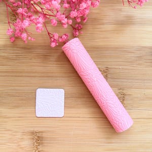 Può includere: Un mattarello rosa e un piccolo timbro quadrato con un motivo floreale. Il mattarello è cilindrico e presenta un motivo in rilievo. Il timbro è quadrato con un motivo floreale in rilievo simile. Fiori rosa sullo sfondo.