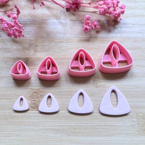 Pode incluir: Cortadores de argila polimérica rosa em vários tamanhos, dispostos sobre uma superfície de madeira. Os cortadores são triangulares com um recorte oval central. Flores rosas adornam o fundo.