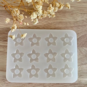 Könnte beinhalten: Eine transparente, rechteckige Silikonform mit zwölf blütenförmigen Vertiefungen. Jede Blume hat fünf Blütenblätter und eine kreisförmige Mitte. Die Form ist für das Basteln bestimmt und liegt auf einer hellen Holzoberfläche.