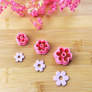 Peut inclure: Des découpoirs à biscuits roses en forme de fleurs et des décorations en fondant en forme de fleurs sont disposés sur une surface en bois. Les découpoirs sont d'un rose vif et les fleurs en fondant sont d'un doux lavande. Des fleurs séchées roses sont en arrière-plan.