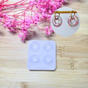 Puede incluir: Un par de pendientes circulares brillantes con aros dorados se muestran junto a un molde de silicona con cuatro cavidades en forma de rosquilla. Los pendientes están llenos de purpurina de colores. Flores rosas en el fondo, todo sobre una superficie de madera.