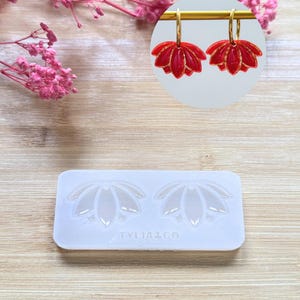 Puede incluir: Pendientes rojos con forma de flor de loto con aros dorados. También se ve un molde de silicona con el texto "TYLIA2CO". Flores rosas en el fondo.