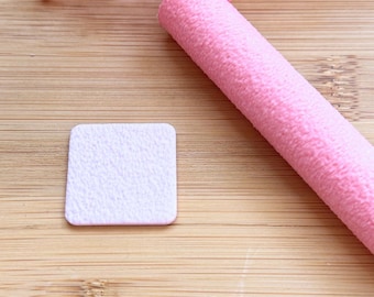 May include: A square, textured, white nail buffer sits next to a pink, cylindrical nail buffer on a wooden surface. Pink floral accents are in the background. The nail buffers are for manicure and pedicure use.