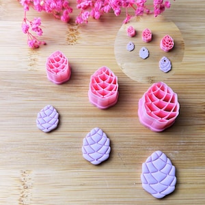 Könnte beinhalten: Eine Sammlung von rosa und lila Tannenzapfen-förmigen Ausstechern und Fondantformen. Die Ausstecher sind in verschiedenen Größen und liegen auf einer Holzoberfläche, mit rosa Blüten im Hintergrund. Die Formen sind aus Kunststoff.