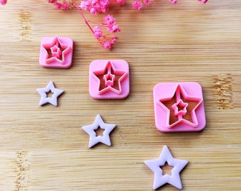 May include: Pink and brown star-shaped cookie cutters and star-shaped cookies are arranged on a wooden surface. The cutters have a square base with a star cutout. Pink flowers are in the background.
