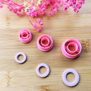 Peut inclure: Ensemble de découpoirs à biscuits roses et violets en forme de donut, de différentes tailles. Les découpoirs sont en plastique et disposés sur une surface en bois. Des fleurs roses sont en arrière-plan.
