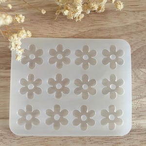 Puede incluir: Un molde de silicona transparente y rectangular con doce cavidades en forma de flor. Cada flor tiene cinco p&eacute;talos y un centro circular. Dise&ntilde;ado para la creaci&oacute;n de elementos decorativos.